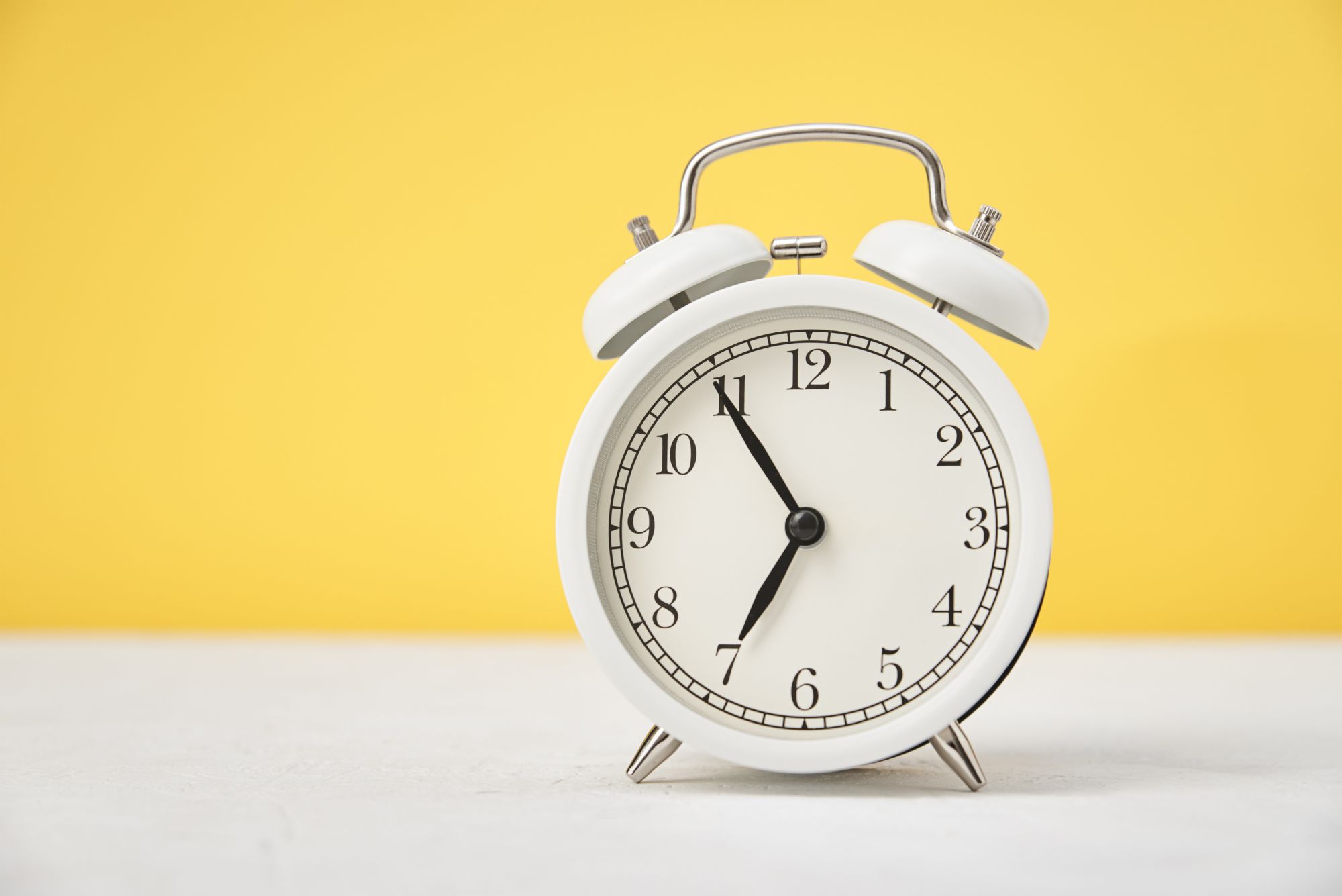 A white analog clock sits on a table against a yellow background